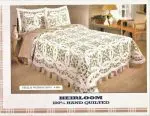 A bedroom features a bed adorned with an Irish Green & White Double Wedding Ring Handmade Quilt, showcasing a beautiful floral pattern. The set is complete with matching pillow shams. A wooden nightstand with a lamp and a chair are positioned next to the bed. Text reads "Heirloom 100% Hand Quilted.