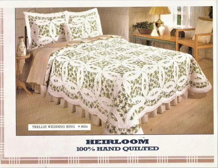 A bedroom features a bed adorned with an Irish Green & White Double Wedding Ring Handmade Quilt, showcasing a beautiful floral pattern. The set is complete with matching pillow shams. A wooden nightstand with a lamp and a chair are positioned next to the bed. Text reads "Heirloom 100% Hand Quilted.