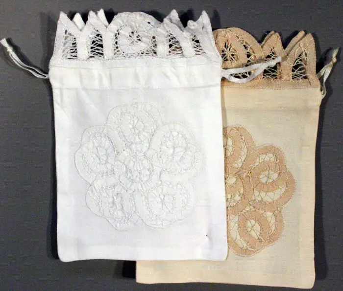 A pair of fabric pouches designed with Battenburg lace, one in white and the other in beige, each featuring intricate lace patterns on the front. Both come with drawstrings and a decorative lace trim at the top.