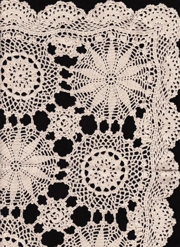 A close-up view of a knitted lace pattern with intricate floral and doily-like designs. The delicate craftsmanship features a repeating motif of circular and star-shaped elements, bordered by a decorative scalloped edge. The lace is off-white or beige against a dark background.