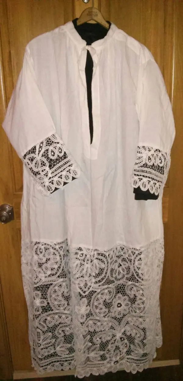 A long white garment with intricate lace detailing on the sleeves and bottom hem is displayed on a hanger. The dress has a round neck with a slit down the front, and the backdrop consists of wooden doors.