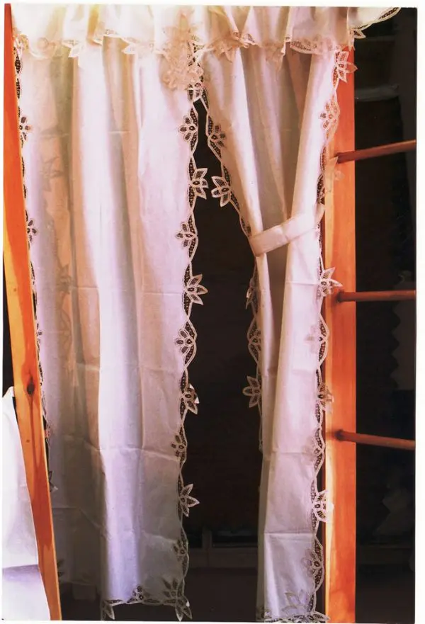 A pair of white lace curtains tied back and hanging from a wooden rod. The curtains feature intricate floral designs along the edges. The setup is in a cozy indoor setting with warm lighting.