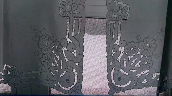 A close-up of a lacy curtain featuring an intricate floral design. The curtain is partially sheer, allowing light to pass through, and two larger floral patterns with leaves and stems are prominently visible. The background beyond the curtain is bright and indistinct.