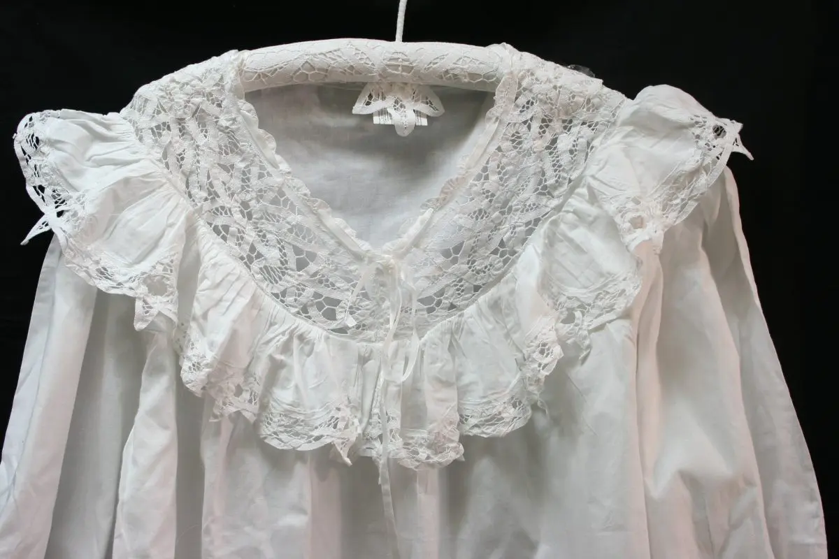 A white blouse with an elaborate lace collar and ruffled edges is displayed on a hanger against a black background. The lace collar features intricate patterns and adds a decorative touch to the garment.