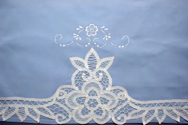 A light blue fabric adorned with intricate white floral lace embroidery at the bottom. The lace features a symmetrical flower and leaf pattern, and there is a smaller embroidered floral design above it. The design is delicate and ornate.