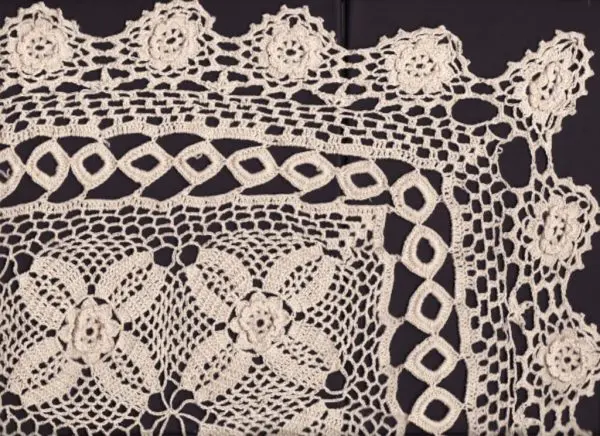 A detailed crochet lace pattern with floral and geometric designs. The intricate lace features interconnected flower motifs and diamond shapes, with a delicate border showcasing a series of small, intricate flowers. The lace is displayed on a dark background.