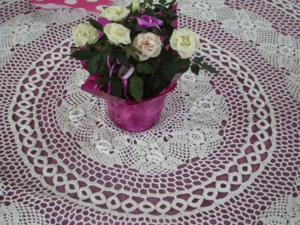 A bouquet of small white roses with green leaves is arranged in a pink pot and placed on a large, intricately crocheted lace doily. The doily features floral and geometric patterns, with a round shape and white thread. The table underneath is purple.
