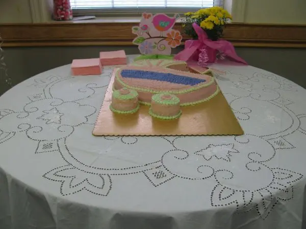 A cake shaped like a baby carriage with pink and green icing is placed on a table covered with a white lace tablecloth. The table also holds pink napkins, a greeting card with a bird design, and a small bouquet of yellow flowers wrapped in pink foil.