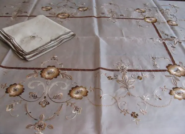 Embroidered tablecloth and napkin set featuring intricate floral designs in beige, brown, and gold tones on a light fabric background. A folded napkin is placed on the top left corner of the tablecloth. The rest of the cloth displays a repeating floral pattern.