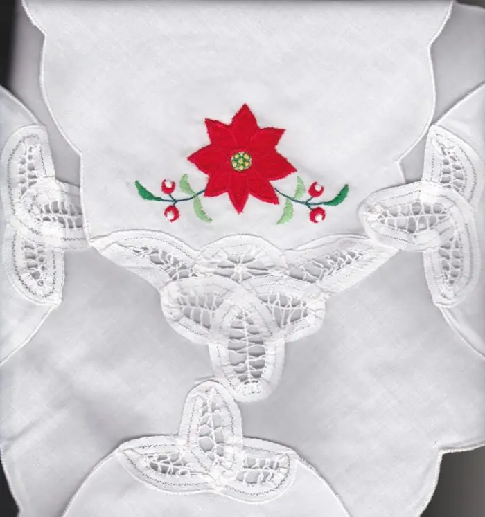 A white fabric with intricate Battenburg lace cutouts features a bright red and green embroidered Poinsettia design. The fabric's design is symmetrical and adds an elegant touch to the piece.