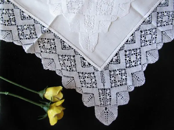 A delicate white lace tablecloth with intricate patterns is displayed against a dark background. Two small yellow roses lie diagonally across the bottom-left corner, adding a touch of color.