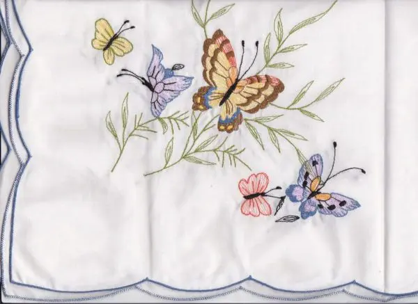 A white fabric with a blue scalloped edge features an embroidered design of colorful butterflies and green leaves. The butterflies are in shades of yellow, purple, orange, pink, and blue, creating a vibrant and delicate pattern.