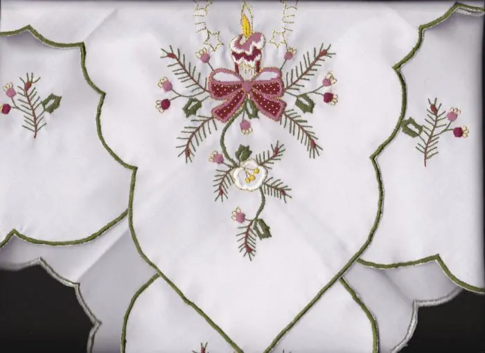 White fabric with green-edged scalloped edges, featuring an embroidered Floral and Holly branches design. The pattern includes a candle, berries, and leaves in shades of red, pink, and green, with a central decorative bow.