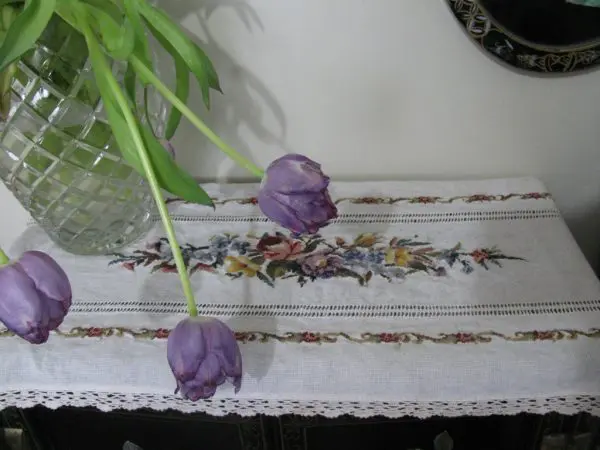 A crystal vase with drooping purple tulips sits on an embroidered floral table runner that is draped over a dark wooden table. The runner features a central design of colorful flowers framed with intricate stitching. A partial view of a decorative mirror is visible.