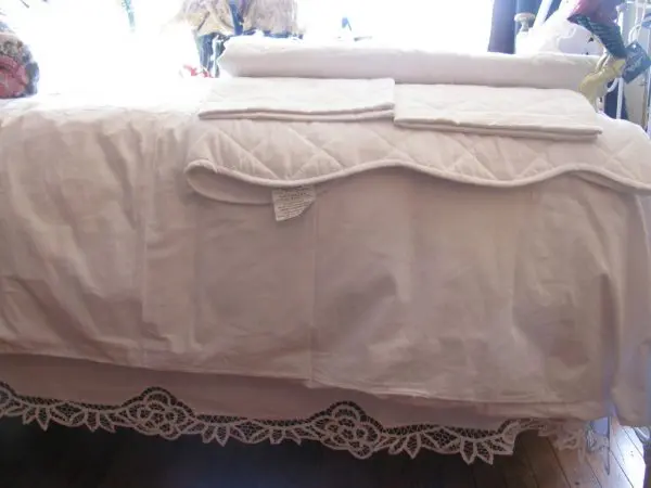 A neatly made bed features a white quilted bedspread with matching pillowcases and a folded quilt. The bed skirt has an intricate lace design that drapes down to the wooden floor. There are additional fabric items and decorative pillows at the head of the bed.