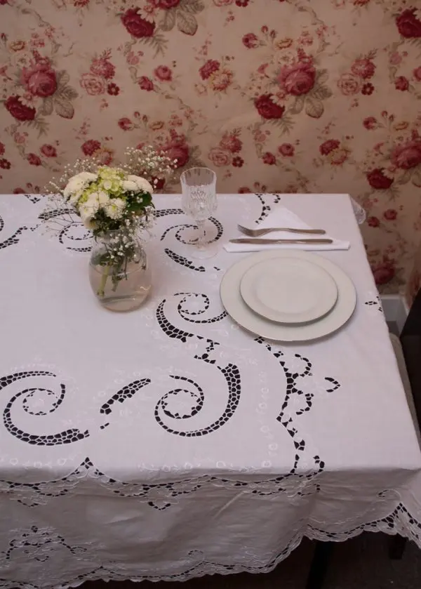 A small table is covered with a white embroidered tablecloth, set with a stack of white plates, a fork and knife on a white napkin, a glass, and a small bouquet of flowers in a jar. The background features wallpaper with a floral pattern.