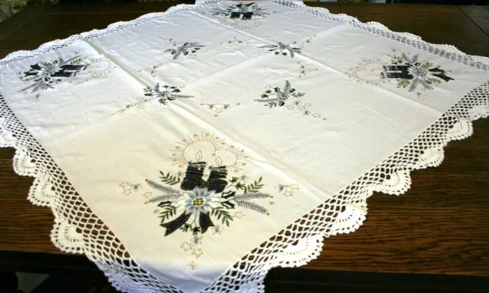 A square tablecloth with intricate embroidery of flowers and candles, featuring delicate lace edges, is spread over a dark wooden table.