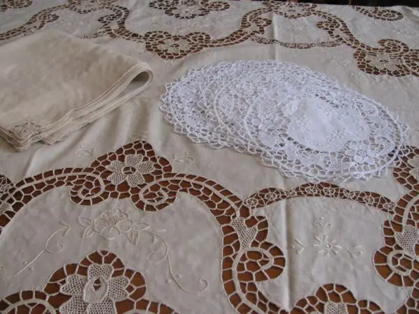 A table is covered with an intricately designed white lace tablecloth featuring floral and geometric patterns. On the table, there is a neatly folded stack of cream-colored cloths and several white lace doilies arranged in a stack beside it.