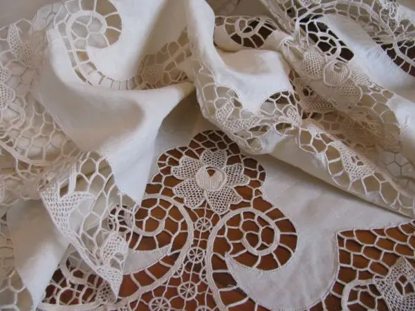 Delicate, cream-colored lace fabric with intricate floral and geometric patterns, partly draped and folded, showcasing fine, detailed craftsmanship. The design features flowers and spirals, creating an elegant and intricate appearance.