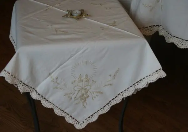 A small table is covered with a white tablecloth adorned with delicate lace trim and intricate embroidered designs. On top of the table is a single small candle in a star-shaped holder. The tablecloth has a lace edge and nature-inspired embroidery in gold thread.