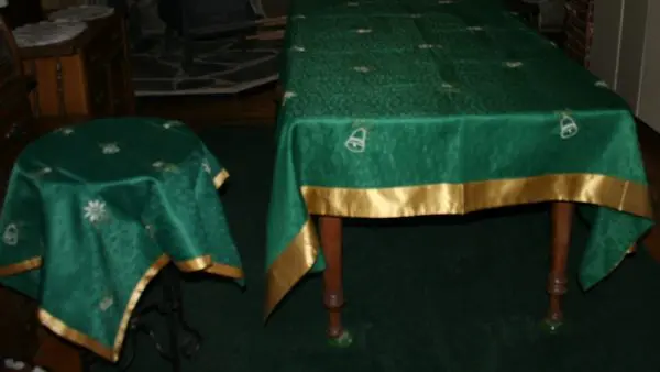 A dining room scene showing a large rectangular table and a small round table, both covered with green tablecloths featuring a gold trim and circular embroidered designs. The background includes wooden furniture and a stone fireplace.