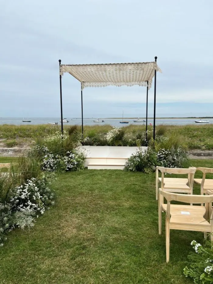 An outdoor wedding setup featuring the unique Wedding Chuppach Canopy, a SnowFlake Crochet Bed Cover overlooks the ocean, with greenery and white flowers enhancing the ambiance. Wooden chairs are meticulously placed on a lush grassy lawn, while a few boats can be seen in the distance beneath a cloudy sky.