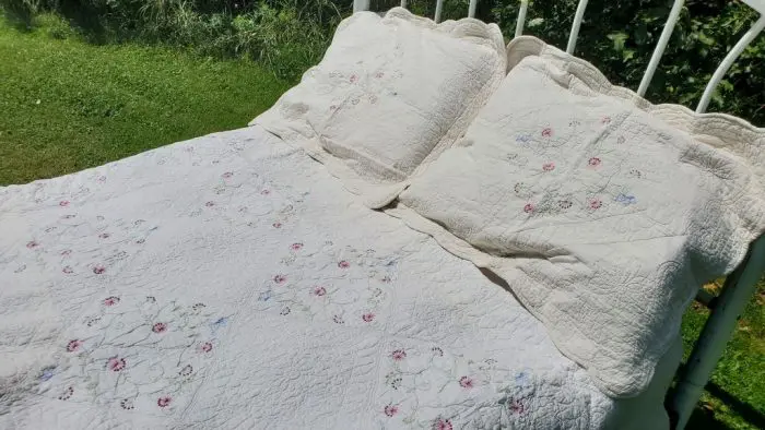 A white metal bed frame stands on grass, adorned with the Fairytale Flower Garden Quilt Cotton Embroidery Whole Cloth and two pillows. The quilt and pillows are intricately embroidered with delicate floral designs in light pink, blue, and green hues. Verdant greenery is visible in the background, suggesting an outdoor setting.