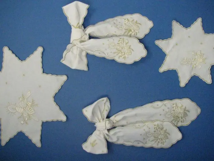 A set of white and gold-embroidered ornaments on a blue background, including two star-shaped pieces and two bow-shaped decorations. The embroidery features floral patterns and delicate details.