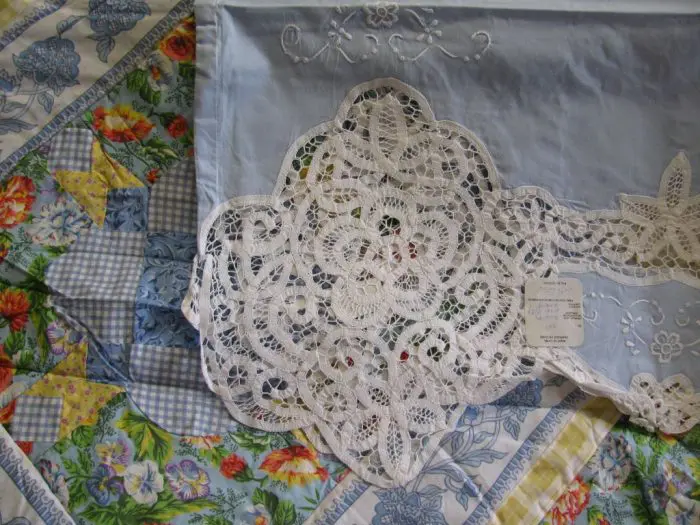 A sky blue embroidered cloth with a delicate white lace edging rests on a 7-piece French Country Quilt, featuring colorful floral and checkered patterns. A tag is attached to the lace piece about the sheet, enhancing the vibrant backdrop.