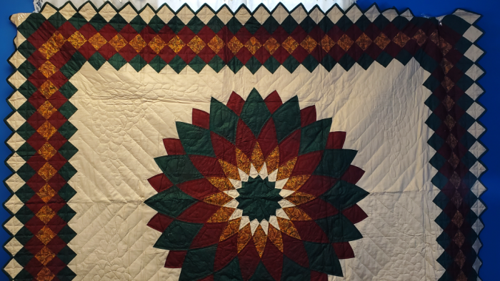 The Nostalgia Brand 1980s Giant Dahlia Handmade Quilt features a central star pattern in green, burgundy, and yellow with intricate diamond borders against a cream background, displayed on a blue wall.