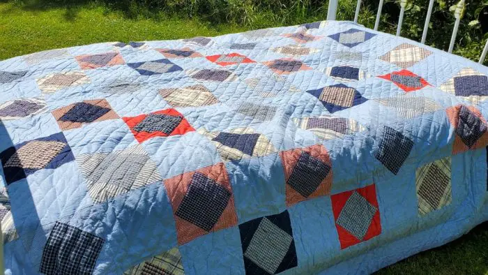 A MUSEUM FLANNEL PLAID PATCHWORK QUILT with ticking fabric backing is draped over a bed outdoors, showcasing its geometric pattern comprised of blue, red, black, and beige squares and triangles. Sunlight softly highlights the textures and colors against the backdrop of green grass.