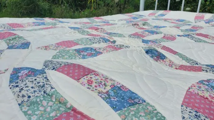 A close-up of the "Courtship Wedding Ring Orange Peel" quilt showcases a patchwork design with various floral patterns in pink, blue, green, and white. The quilt is spread out on a bed with a metal frame, and lush greenery can be seen in the background. Hand quilting featured Orange Peel design.