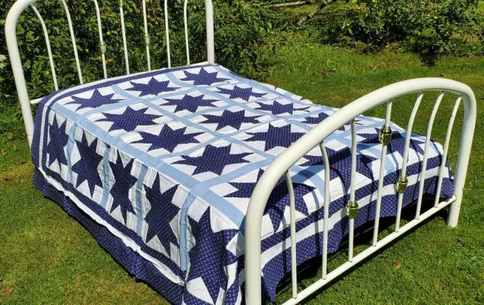 A white metal bed frame sits on a grassy lawn, adorned with the Blue Paisley Morning Star Quilt, featuring large blue stars and squares on a white and light blue background. Lush green plants and a stone wall surround the bed.