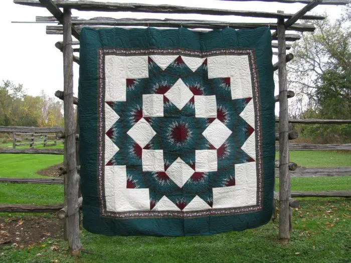 The Nostalgia 1980s Donna Sharp Broken Star Original Quilt with a multicolored geometric star pattern featuring dark green, ivory, and maroon colors is displayed on a rustic wooden frame against grass and trees.