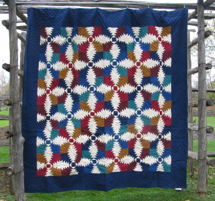 The Flannel Pineapple Hand Quilted 100% Cotton-Jewel Tones quilt, showcasing intricate geometric patterns in red, blue, brown, and ivory shapes resembling pine cones on a deep blue border, hangs on a wooden frame outdoors over grass.