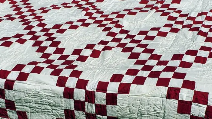 The Irish Chain Quilt in burgundy features a red and white checkerboard pattern with intersecting diagonal lines, creating a lattice-like look that adds a traditional and cozy feel to the bedding.