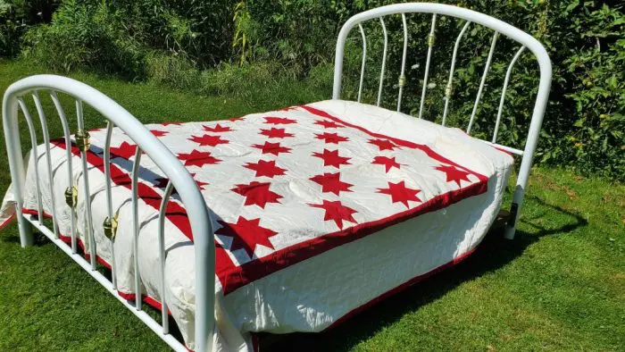 A white metal bed frame is set outdoors on green grass, covered with "The burgundy RED Star Quilt Simple Red & White Cotton." Bushes are visible in the background.