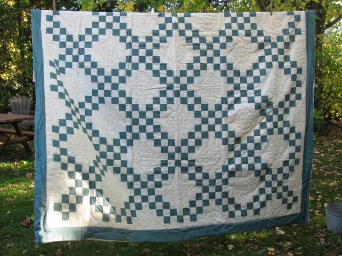 The Double Irish Chain Handmade Quilt - Antique Green is elegantly displayed outdoors, showcasing an intricate pattern of interlocking teal squares on a ivory background. It hangs from a line, amidst lush green foliage with a wooden chair in the backdrop.