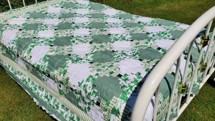 Set outdoors on grass, the bed features the Sparkling Green & White Courthouse Steps Prairie Point Border quilt with floral and geometric patterns. The bed has a white metal frame adorned with brass accents.