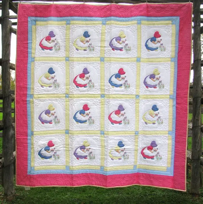 The Sun Bonnet Sue or Little Dutch Girl Hand Appliqué Quilt hangs on a wooden frame, displaying a repeated pattern of a child in a hat kneeling beside flowers. It features a grid design with brightly-colored borders in pink, yellow, and blue squares.