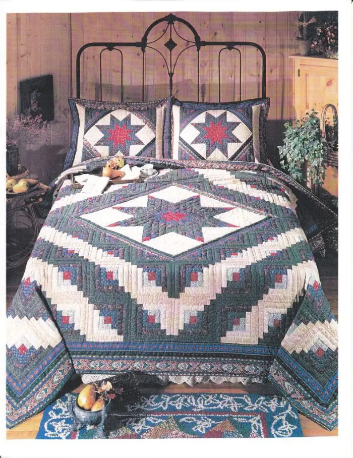 A cozy bedroom showcases a metal-frame bed adorned with the Log Cabin Lone Star 1980 American Pacific Design quilt and pillows in red, blue, and white star patterns. The room features a decorative rug, potted plant, and bedside table with cheerful plush toys.