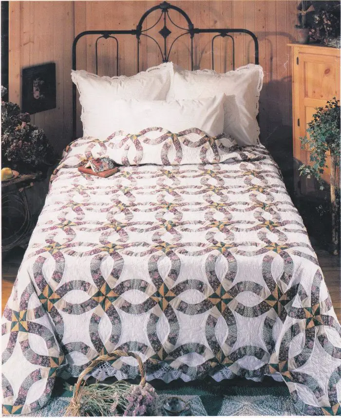 A bed with a Purple & Green 1980s Double Wedding Ring Quilt on an ornate metal headboard, set against wooden paneled walls. The room includes white pillows, a wooden cabinet, and decorative plants, welcoming ambiance of rustic charm