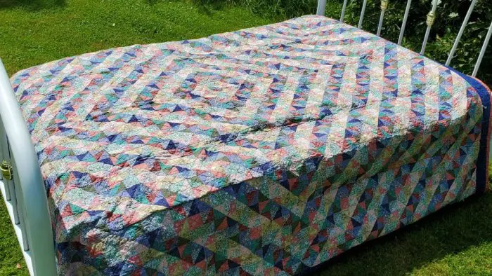 The Broken Dish Rare Trip Around the World Quilt, showcasing a multicolored geometric design with blue, green, pink, and white triangles forming diamonds, rests on a white metal bed frame outdoors. Grass surrounds the setup.