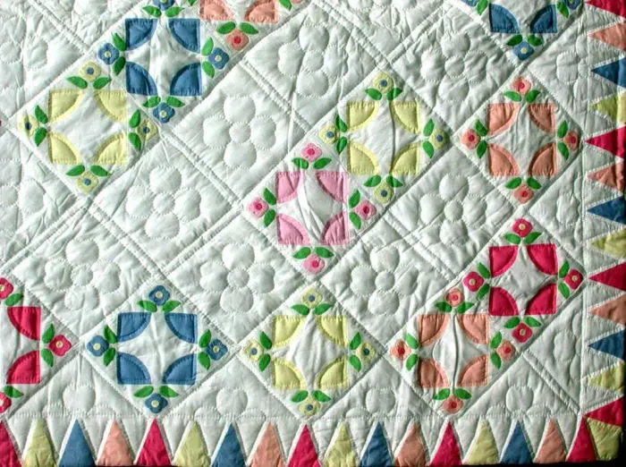 The Sunshine Cathedral Window Ice Cream Colours Hand-Quilted Quilt showcases a vibrant, geometric design on a white background with diamond patterns in blue, green, pink, and orange. The quilt's edges are adorned with pink and blue triangular shapes featuring intricate quilting and floral motifs.