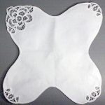 An Elite Battenburg Lace Bun Warmer Cover in ecru cotton, featuring lace embroidery on one corner and smaller lace accents on two corners, displayed against a gray background.