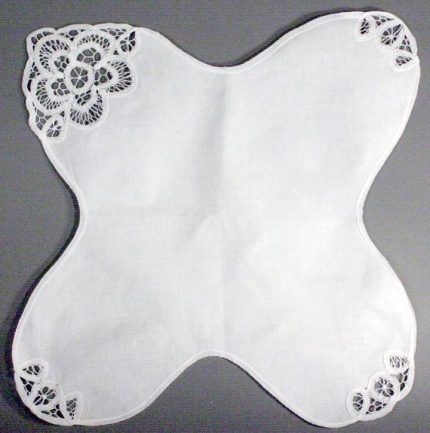 An Elite Battenburg Lace Bun Warmer Cover in ecru cotton, featuring lace embroidery on one corner and smaller lace accents on two corners, displayed against a gray background.