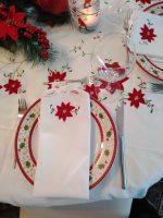 A festive table setting is highlighted by the XMas Dinner Tablecloth, which showcases red poinsettia designs and Battenburg lace on a white cotton oblong. A matching white napkin with a poinsettia design is elegantly placed on top of a decorative plate. Completing the arrangement are a glass, fork, and knife positioned neatly, with a candle and greenery decoration adding to the ambiance.