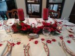 A festive dining table set for a holiday meal is adorned with the XMas Dinner Tablecloth featuring a Poinsettia and Battenburg Lace design in vibrant reds and greens. The table displays white napkins, red candles, elegant glassware, and a centerpiece with red flowers and greenery to complete the ambiance.