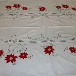 The XMas Dinner Tablecloth Poinsettia & Battenburg Lace Cotton Oblong showcases a white base adorned with red embroidered poinsettias accompanied by green leaves. A finely crafted Battenburg lace strip, featuring intricate patterns, runs horizontally across the center. The symmetrical design places poinsettias on both sides of the lace strip, creating an elegant holiday look.