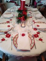 A festive dining table set for Christmas, featuring the XMas Dinner Tablecloth with a poinsettia and Battenburg lace design, complemented by red and white plates, wine glasses, white napkins, and a red candle centerpiece surrounded by greenery and decorations.